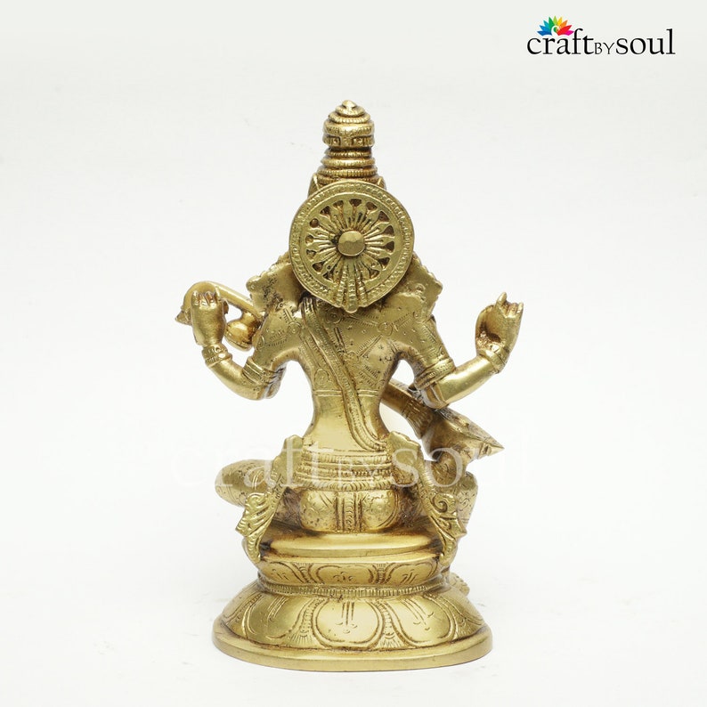 Puede incluir: Estatua de lat&oacute;n de una deidad, vista desde atr&aacute;s, con detalles intrincados. La estatua presenta un dise&ntilde;o de rueda en forma de halo y est&aacute; sentada sobre una base de flor de loto. La estatua es de color dorado y sobre un fondo blanco.