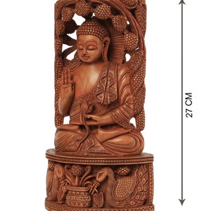 Op de afbeelding: Gedetailleerd houten Boeddha-beeld, 27 cm hoog, met ingewikkelde gravures. Het beeld zit op een decoratieve basis, 12 cm breed en 5 cm diep, met pauwen- en bloemmotieven. De tekst "craft by soul" is inbegrepen.
