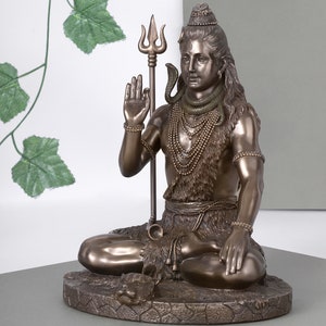 Puede incluir: Estatua de bronce de una deidad sentada, adornada con collares en capas y una serpiente. La figura sostiene un tridente y tiene el pelo largo. La estatua est&aacute; sobre una base texturizada.