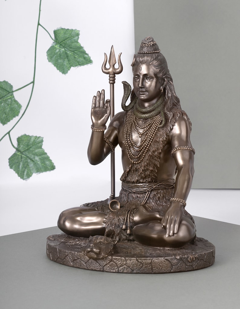 Puede incluir: Estatua de bronce de una deidad sentada, adornada con collares en capas y una serpiente. La figura sostiene un tridente y tiene el pelo largo. La estatua est&aacute; sobre una base texturizada.