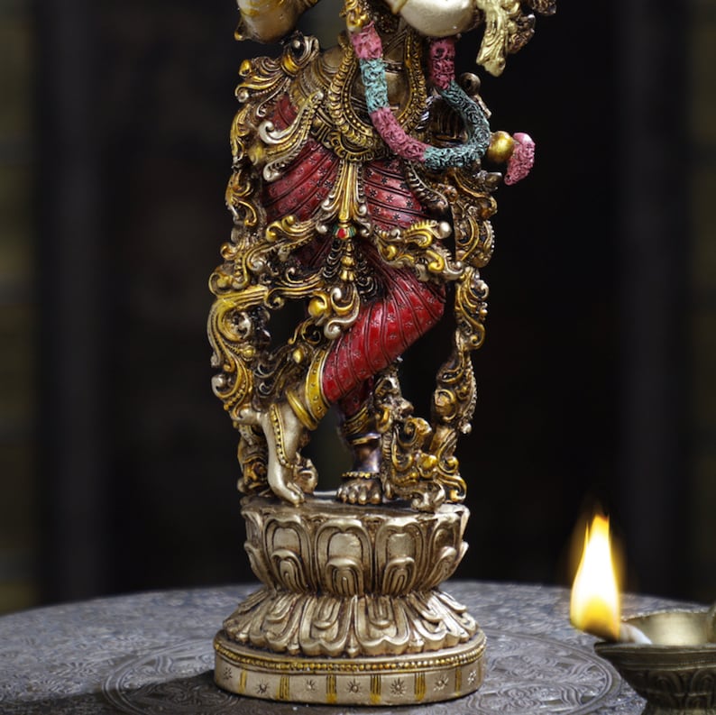 May include: Ornate statue of a deity, featuring intricate gold and red details. The figure stands on a lotus flower base. A colorful garland and elaborate jewelry adorn the statue. The statue is captured in a close-up shot.