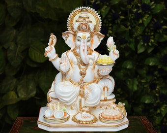 May include: A white and gold statue of the Hindu deity Ganesha, seated with multiple arms, holding various items. The statue features intricate details, including a crown and ornate jewelry. The background is a blurred green.