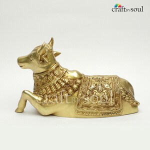 May include: A golden brass sculpture of a reclining cow, adorned with intricate embossed patterns on its body and a decorative blanket. The sculpture has detailed horns and ears, and the Craft by Soul logo is visible in the upper right corner.