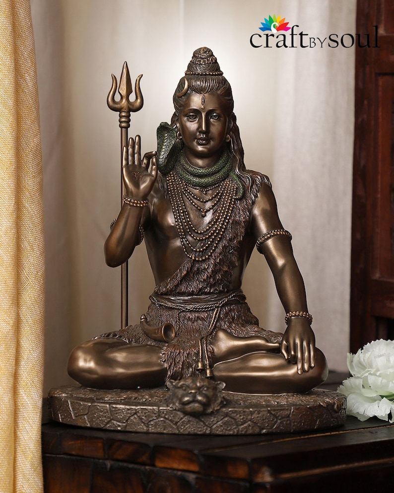 Puede incluir: Estatua de color bronce de una deidad sentada, sosteniendo un tridente y adornada con collares y una serpiente. La figura tiene una expresi&oacute;n serena y est&aacute; sentada sobre una base circular detallada. Es un art&iacute;culo religioso o espiritual.