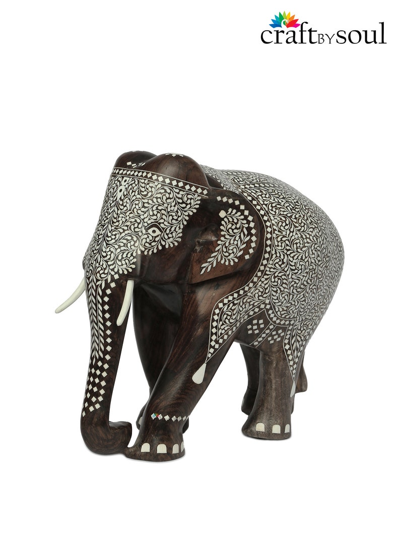 Puede incluir: Una escultura de elefante de madera hecha a mano con intrincados detalles de incrustaciones blancas. El elefante marr&oacute;n oscuro presenta colmillos y est&aacute; decorado con patrones florales y geom&eacute;tricos. La escultura es un objeto decorativo.