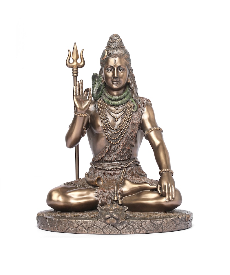 Puede incluir: Estatua de bronce de una deidad sentada, sosteniendo un tridente y adornada con una serpiente y collares en capas. La figura est&aacute; sentada en una pose meditativa sobre una base texturizada, con detalles intrincados en la ropa y las joyas.