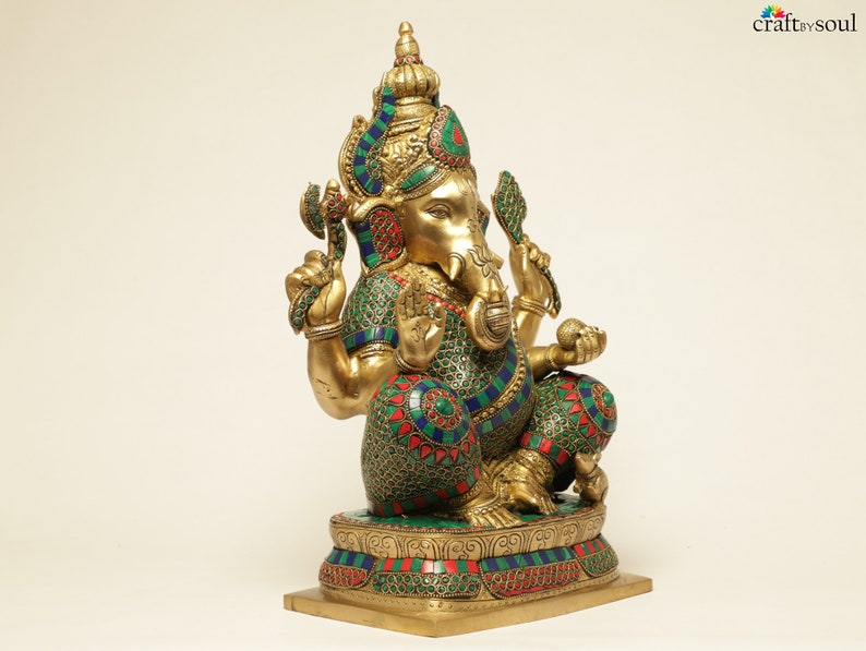 May include: A gold-colored statue of the Hindu deity Ganesha, adorned with colorful accents of green, red, and blue. The statue is intricately detailed, depicting Ganesha in a seated position with multiple arms and a large head. The base is rectangular.