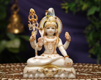 May include: A white and gold statue of a deity in a meditative pose. The statue has multiple arms and is decorated with intricate details, including a serpent and a halo. The figure holds a trident against a blurred background.