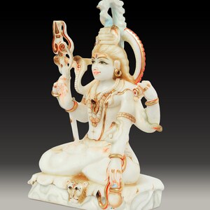 May include: A white and gold statue of a deity, seated in a meditative pose. The statue features multiple arms, intricate jewelry, and a serpent coiled around the neck. The base has a lion's head carved into it. The statue is set against a dark background.