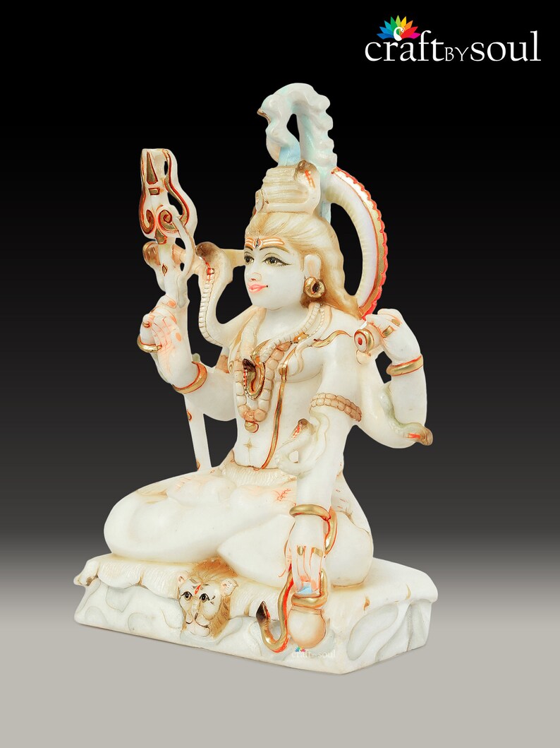May include: A white and gold statue of a deity, seated in a meditative pose. The statue features multiple arms, intricate jewelry, and a serpent coiled around the neck. The base has a lion's head carved into it. The statue is set against a dark background.