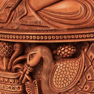 Op de afbeelding: Close-up van een gedetailleerde houten snijwerk. Het snijwerk toont een zwanenachtige vogel die een fluit bespeelt, ingewikkelde patronen en een zittende figuur. Het hout heeft een warme, natuurlijke tint, die het vakmanschap laat zien.