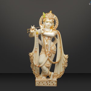 Puede incluir: Estatua blanca y dorada de una deidad tocando la flauta. La estatua tiene detalles intrincados, incluyendo un halo, joyas y ropa drapeada. La base est&aacute; decorada con un patr&oacute;n repetido. El fondo es un degradado de gris.