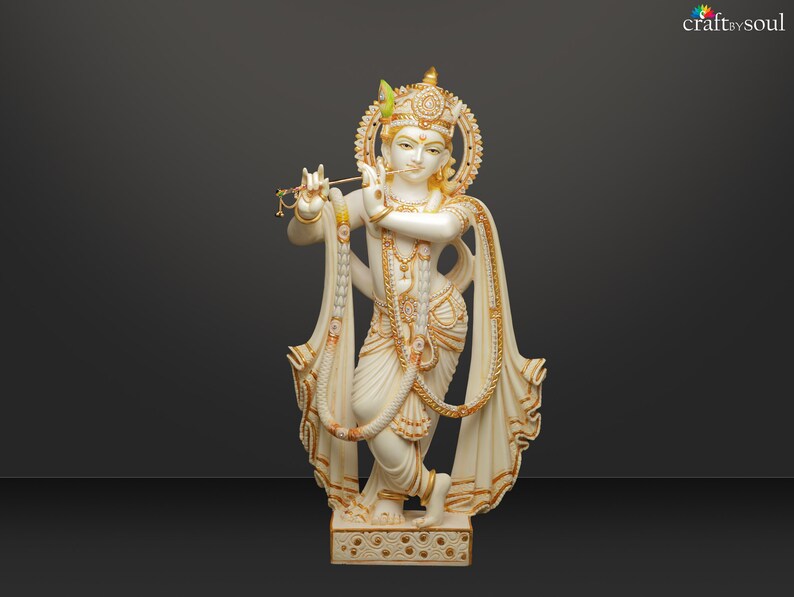 Puede incluir: Estatua blanca y dorada de una deidad tocando la flauta. La estatua tiene detalles intrincados, incluyendo un halo, joyas y ropa drapeada. La base est&aacute; decorada con un patr&oacute;n repetido. El fondo es un degradado de gris.