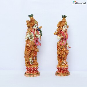 May include: Two ornate statues of deities, one playing a flute and the other standing. Both figures are adorned with gold, red, and green details, including elaborate headdresses and floral garlands. They stand on decorative red and gold bases.