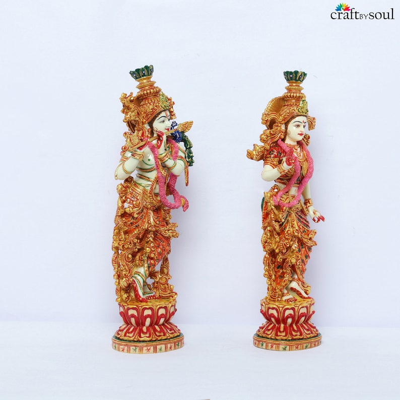 May include: Two ornate statues of deities, one playing a flute and the other standing. Both figures are adorned with gold, red, and green details, including elaborate headdresses and floral garlands. They stand on decorative red and gold bases.