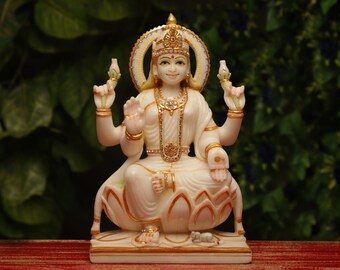 May include: A white and gold statue of a seated deity with multiple arms, adorned with a jeweled crown and necklace. The statue is seated on a lotus flower base, with intricate details and a serene expression. The background is a blurred green.