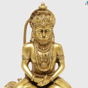 K&ouml;nnte beinhalten: Eine detaillierte Messingstatue einer sitzenden Gottheit mit affen&auml;hnlichem Gesicht, geschm&uuml;ckt mit aufwendigem Schmuck und einer Krone. Die Statue hat einen goldenen Farbton und ist vor einem schlichten Hintergrund platziert. Es ist ein religi&ouml;ses oder spirituelles Objekt.