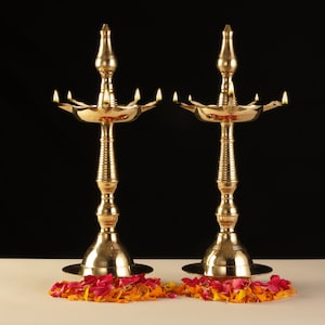 May include: Two ornate brass oil lamps with lit wicks, set against a black backdrop. The lamps have a tall, slender design with a flared base and a multi-tiered top. Red and orange flower petals are scattered at the base of each lamp.