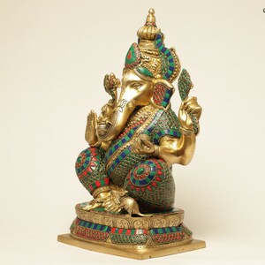 May include: A gold-colored statue of the Hindu deity Ganesha, adorned with colorful accents of green, red, and blue. The statue is intricately detailed, featuring a crown, jewelry, and a mouse at its feet. The base is also decorated with colorful stones.