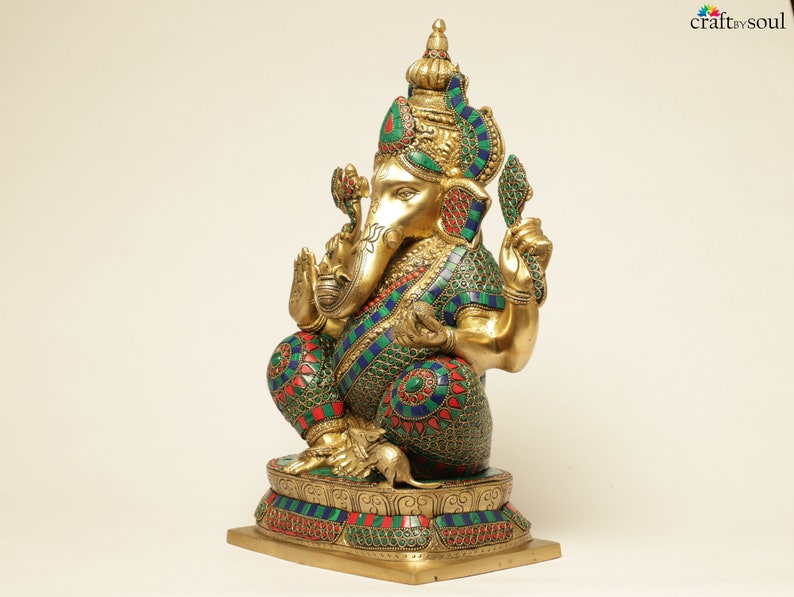 May include: A gold-colored statue of the Hindu deity Ganesha, adorned with colorful accents of green, red, and blue. The statue is intricately detailed, featuring a crown, jewelry, and a mouse at its feet. The base is also decorated with colorful stones.