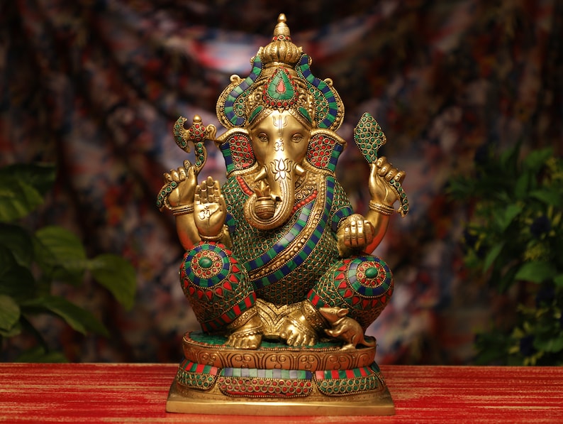 May include: A gold-colored statue of the Hindu deity Ganesha, seated with multiple arms and an elephant head. The statue is adorned with colorful mosaic-like patterns in green, blue, and red. The base is also decorated with similar mosaic designs.