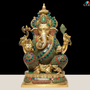 May include: A detailed brass statue of the Hindu deity Ganesha, seated with multiple arms and an elephant head. The statue is adorned with colorful gemstones and intricate patterns, set against a black background. The base is decorated with floral designs.
