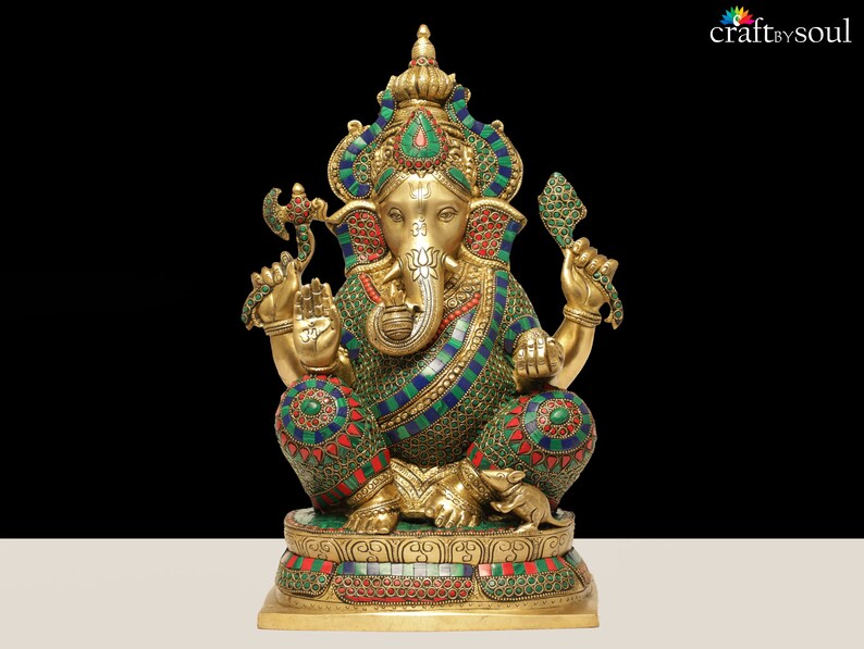 May include: A detailed brass statue of the Hindu deity Ganesha, seated with multiple arms and an elephant head. The statue is adorned with colorful gemstones and intricate patterns, set against a black background. The base is decorated with floral designs.