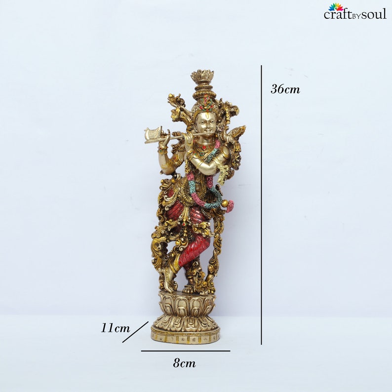 May include: A detailed brass statue of a deity playing a flute, adorned with colorful clothing and jewelry. The statue is 36 cm tall, 11 cm wide, and 8 cm deep. The base is intricately designed, and the figure has a serene expression.