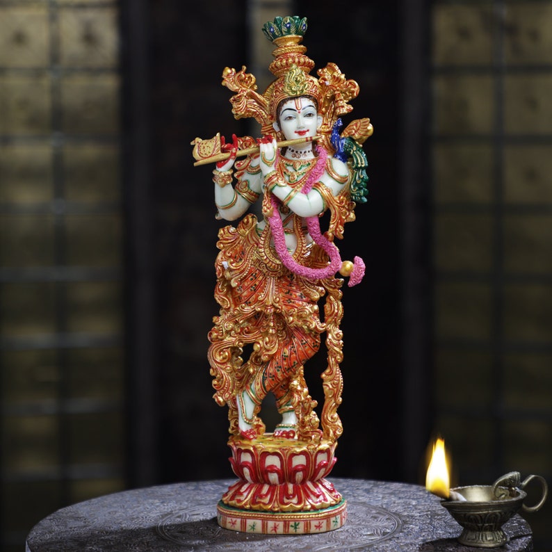 May include: A detailed statue of a deity, featuring a white complexion, ornate gold and red clothing, and a pink garland. The figure is playing a flute. The statue is on a red and gold lotus base.