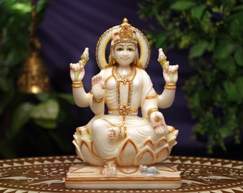 May include: A white and gold statue of a seated deity, adorned with a crown and jewelry. The figure is seated on a lotus flower base, with multiple arms and a halo. The statue has a serene expression and is set against a blurred background.