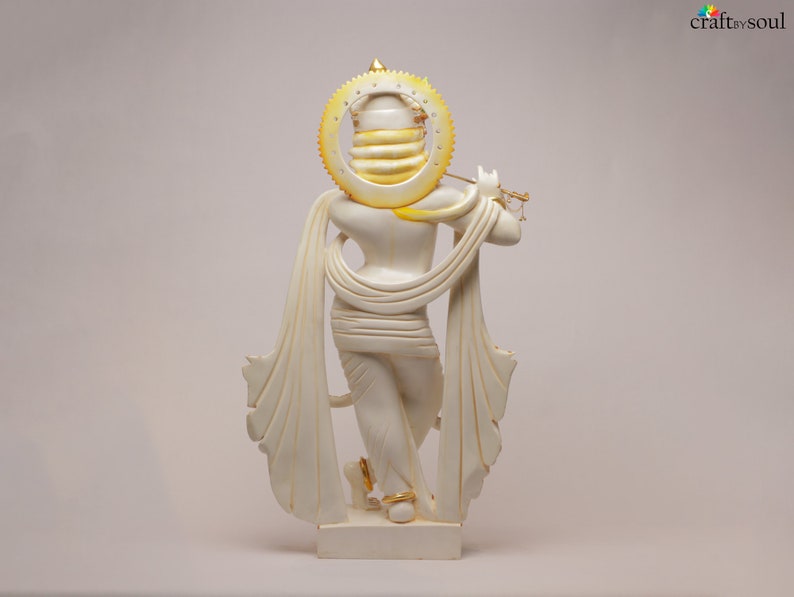 Puede incluir: Estatua color crema de una figura tocando la flauta, con un halo y acentos dorados. La figura est&aacute; envuelta en tela fluida, y la estatua tiene una base rectangular. El dise&ntilde;o general es detallado y elegante.