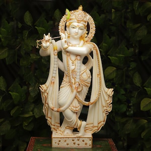 Puede incluir: Estatua blanca y dorada de una deidad tocando la flauta. La estatua tiene detalles intrincados, incluyendo un halo, ropa ornamentada y joyas. El fondo es un exuberante follaje verde.