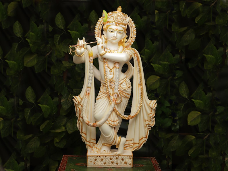 Puede incluir: Estatua blanca y dorada de una deidad tocando la flauta. La estatua tiene detalles intrincados, incluyendo un halo, ropa ornamentada y joyas. El fondo es un exuberante follaje verde.