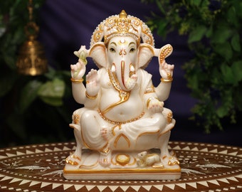 May include: A white and gold statue of the Hindu deity Ganesha, seated in a meditative pose. The statue features intricate details, including a crown, jewelry, and a mouse at the base. The figure is set against a blurred background of greenery.