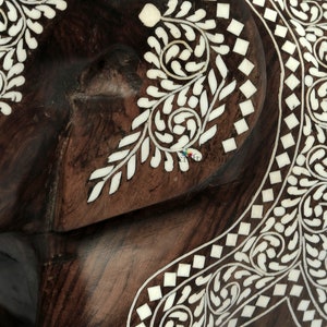 Puede incluir: Primer plano de un objeto de madera marr&oacute;n oscuro con intrincados dise&ntilde;os de incrustaciones blancas. La incrustaci&oacute;n presenta patrones florales y geom&eacute;tricos, creando una superficie decorativa y detallada. La veta de la madera es visible.