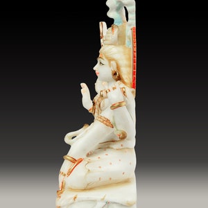 May include: A white marble statue of a seated deity with gold accents and intricate details. The figure has a serene expression, adorned with jewelry and a crown. The statue is set against a dark background, highlighting the craftsmanship.