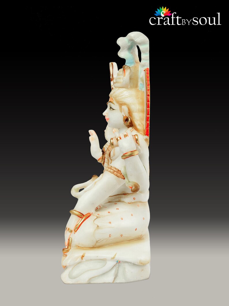 May include: A white marble statue of a seated deity with gold accents and intricate details. The figure has a serene expression, adorned with jewelry and a crown. The statue is set against a dark background, highlighting the craftsmanship.
