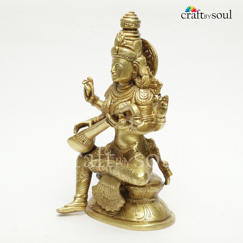 Puede incluir: Estatua de lat&oacute;n de una deidad sentada, adornada con detalles intrincados. La figura sostiene un instrumento de cuerda y est&aacute; sentada sobre una base decorativa. La estatua tiene m&uacute;ltiples brazos y lleva joyas ornamentadas. El color general es dorado c&aacute;lido.