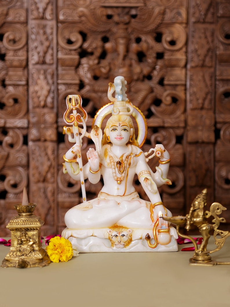 May include: A white and gold statue of a deity with multiple arms, seated in a meditative pose. The statue has intricate details and is adorned with gold accents. A yellow flower and brass objects are in the foreground. The background features a carved wooden panel.