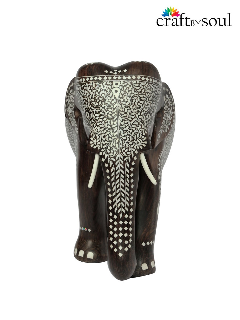 Puede incluir: Una figura de elefante de madera marr&oacute;n oscuro con intrincados patrones de incrustaciones blancas. El elefante tiene colmillos de color marfil y tallas detalladas en la cabeza y el cuerpo. Es un objeto decorativo.