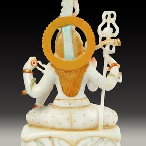 May include: A white marble statue of a deity, viewed from the back. The statue features intricate details, including a golden halo, ornate jewelry, and a staff. The figure is seated in a meditative pose, with a serene expression. The statue is on a decorative base.
