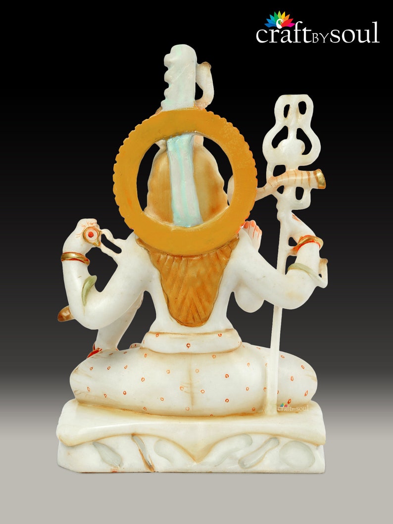 May include: A white marble statue of a deity, viewed from the back. The statue features intricate details, including a golden halo, ornate jewelry, and a staff. The figure is seated in a meditative pose, with a serene expression. The statue is on a decorative base.