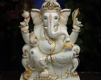 Op de afbeelding: Een wit en goud beeld van de hindoeïstische godheid Ganesha, zittend op een troon. Het beeld heeft ingewikkelde details, waaronder een kroon, sieraden en een slurf met een zoete lekkernij. De basis is rechthoekig en het algehele ontwerp is sierlijk.