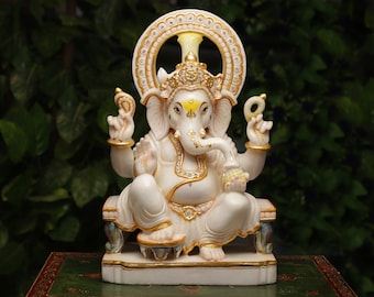 May include: A white and gold statue of the Hindu deity Ganesha, seated in a meditative pose. The statue features intricate details, including a large head, multiple arms, and a decorative halo. The statue is adorned with gold accents and jewels.
