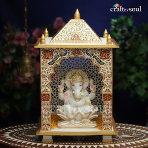 以下が含まれることがあります： 金色のアクセントと複雑な花と孔雀のデザインが施された白い大理石の寺院。蓮の花の上に座るガネーシャの白い像が収められています。構造は格子細工のデザインが特徴です。背景は濃い青色で、寸法はcmです。