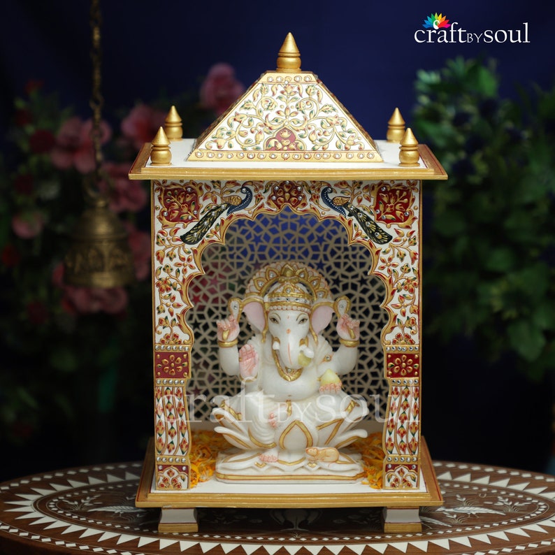 以下が含まれることがあります： 金色のアクセントと複雑な花と孔雀のデザインが施された白い大理石の寺院。蓮の花の上に座るガネーシャの白い像が収められています。構造は格子細工のデザインが特徴です。背景は濃い青色で、寸法はcmです。