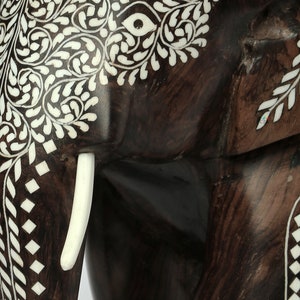 Puede incluir: Primer plano de una escultura de elefante de madera marr&oacute;n oscuro con intrincados dise&ntilde;os de incrustaciones blancas. El cuerpo del elefante presenta patrones florales y geom&eacute;tricos. Se ve un colmillo blanco, que a&ntilde;ade detalle a la obra artesanal.