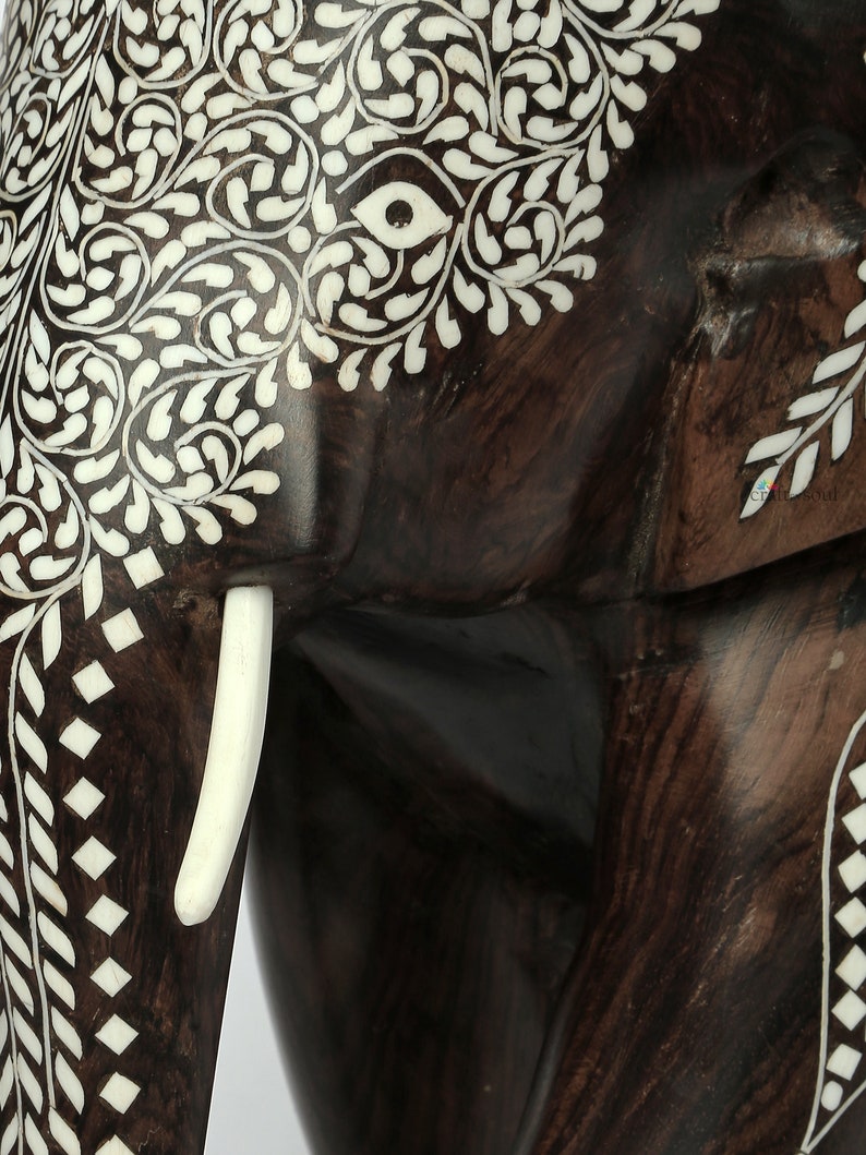 Puede incluir: Primer plano de una escultura de elefante de madera marr&oacute;n oscuro con intrincados dise&ntilde;os de incrustaciones blancas. El cuerpo del elefante presenta patrones florales y geom&eacute;tricos. Se ve un colmillo blanco, que a&ntilde;ade detalle a la obra artesanal.