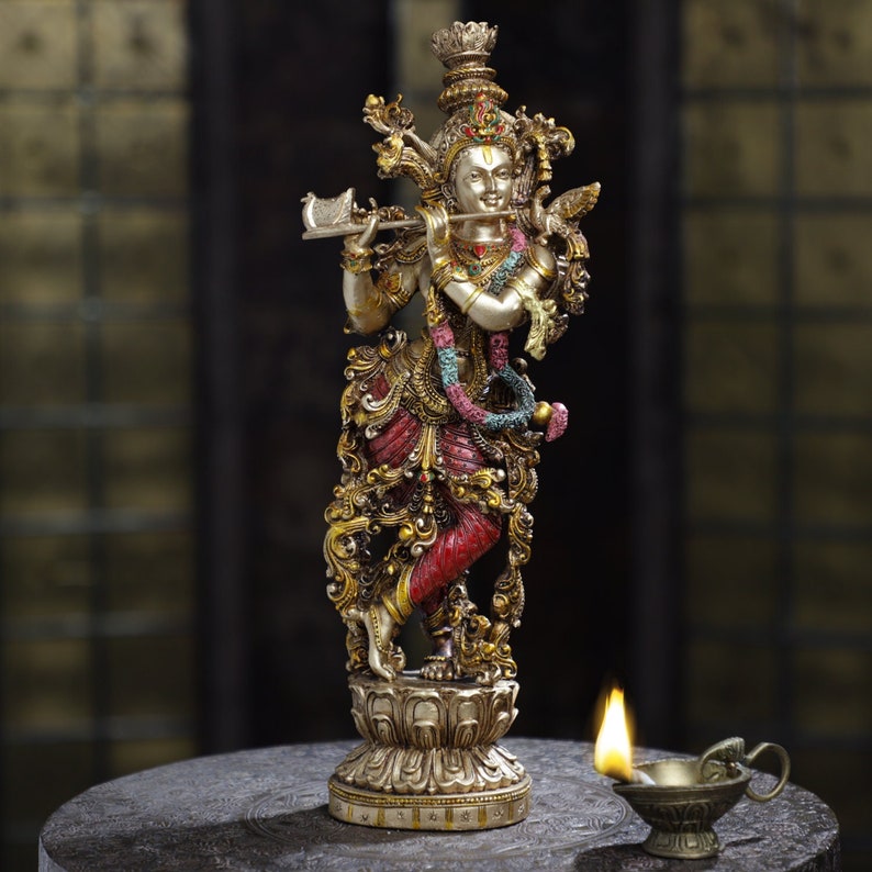 May include: A detailed statue of a deity playing a flute, adorned with intricate gold and red details. The figure stands on a lotus flower base. A small oil lamp with a lit flame is in the foreground.