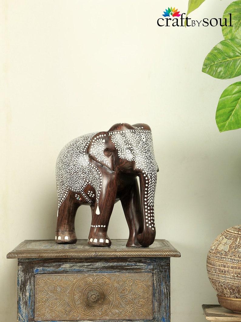 Puede incluir: Una figura de elefante de madera marr&oacute;n oscuro con patrones florales y de puntos blancos. El elefante est&aacute; sobre una peque&ntilde;a mesa ornamentada. El logotipo de Craft by Soul es visible en la esquina superior derecha.